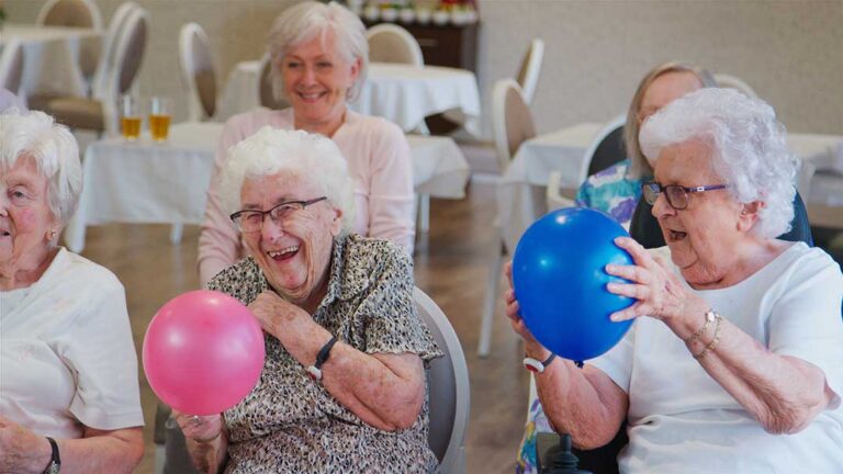 Older adults seated together making seasonal crafts near a bright window with plants and art supplies.